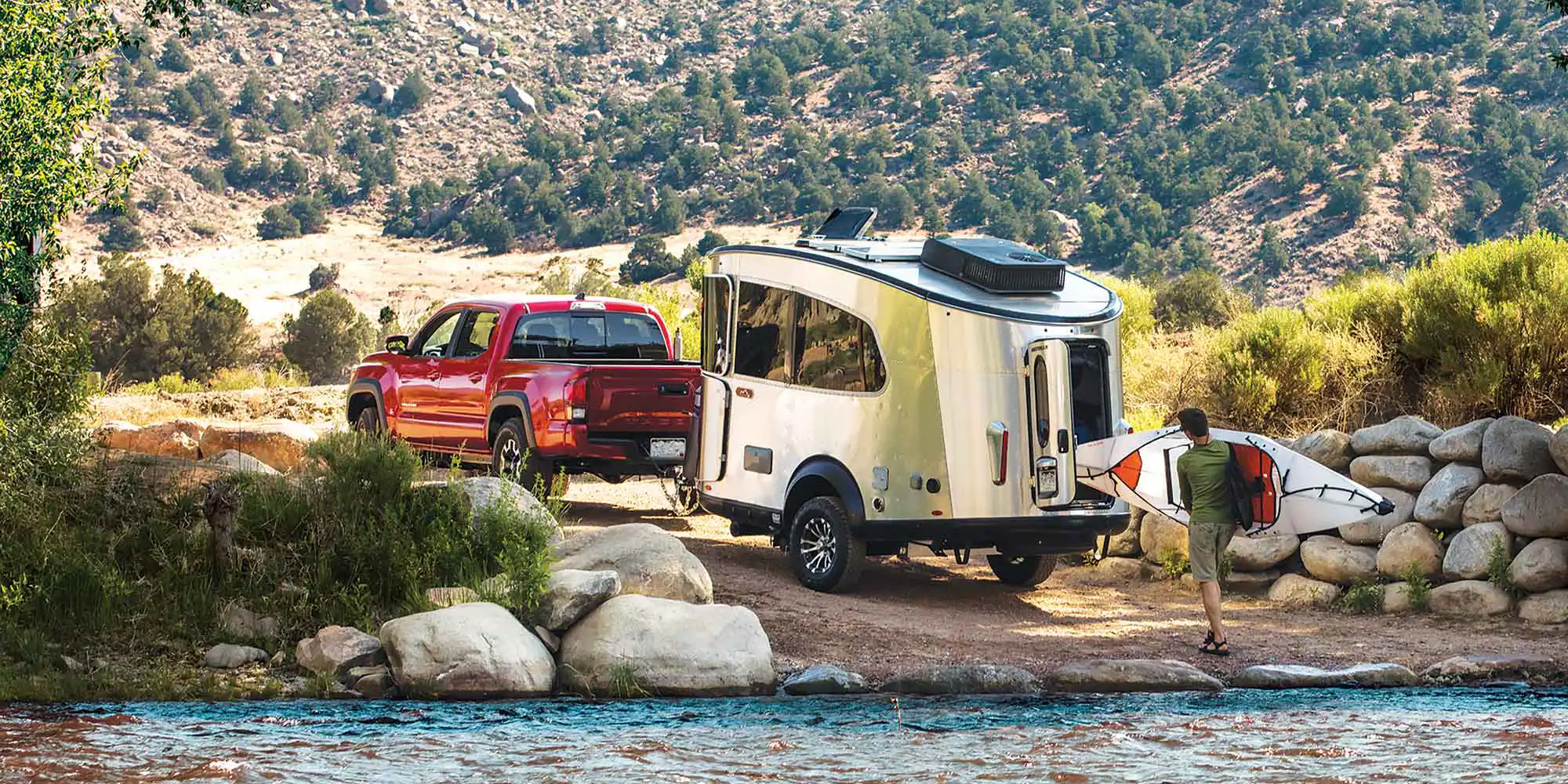 An Airstream Basecamp towed by a pickup at a rocky riverside, a paddler carrying a kayak down to the water