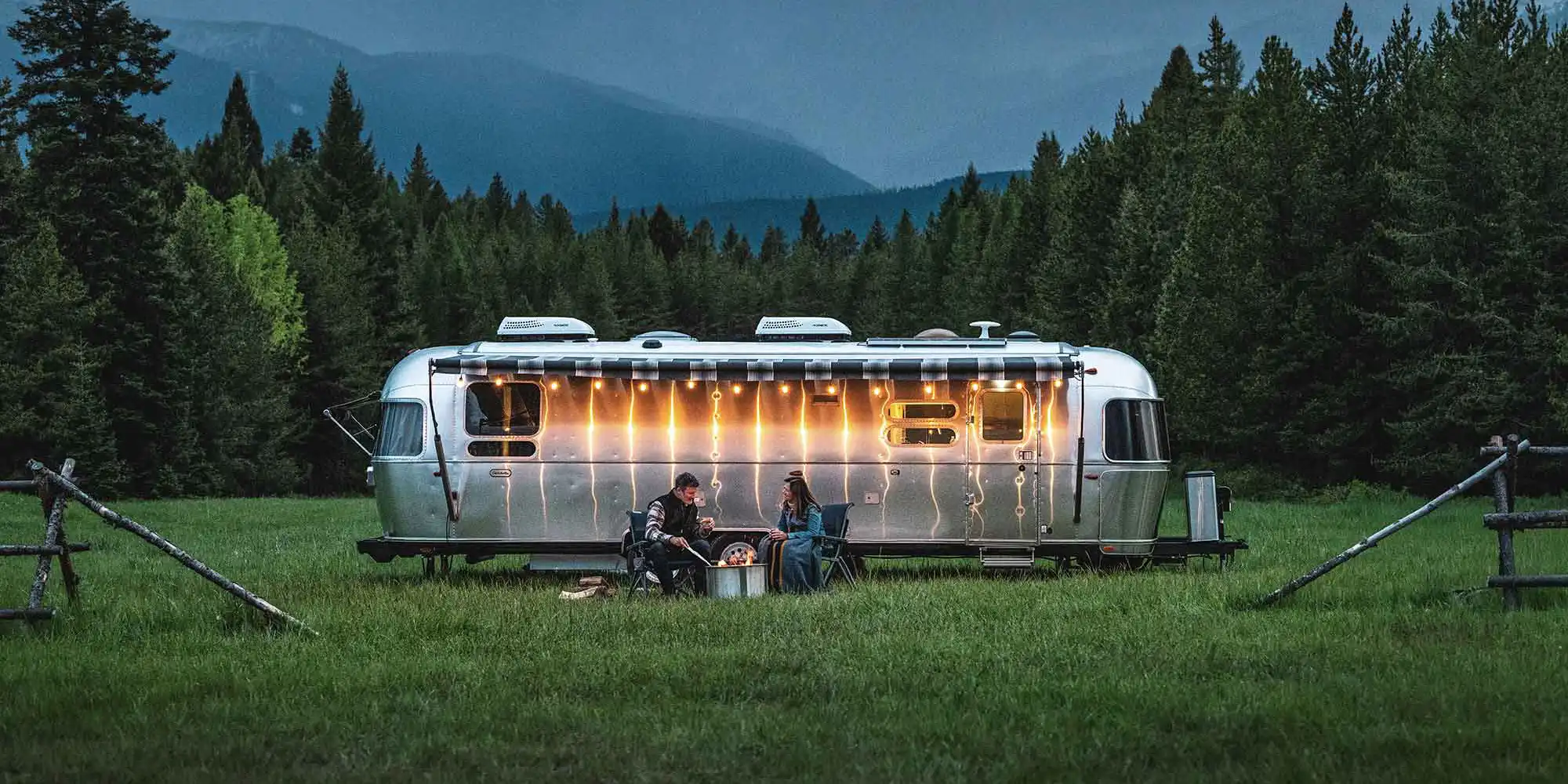 An Airstream travel trailer in a mountain meadow at dusk, string lights along the awning and two people at a fire pit