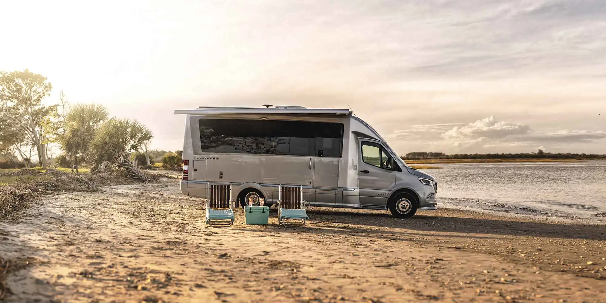 An Airstream Atlas touring coach parked on a beach at sunset with camp chairs and a cooler set out